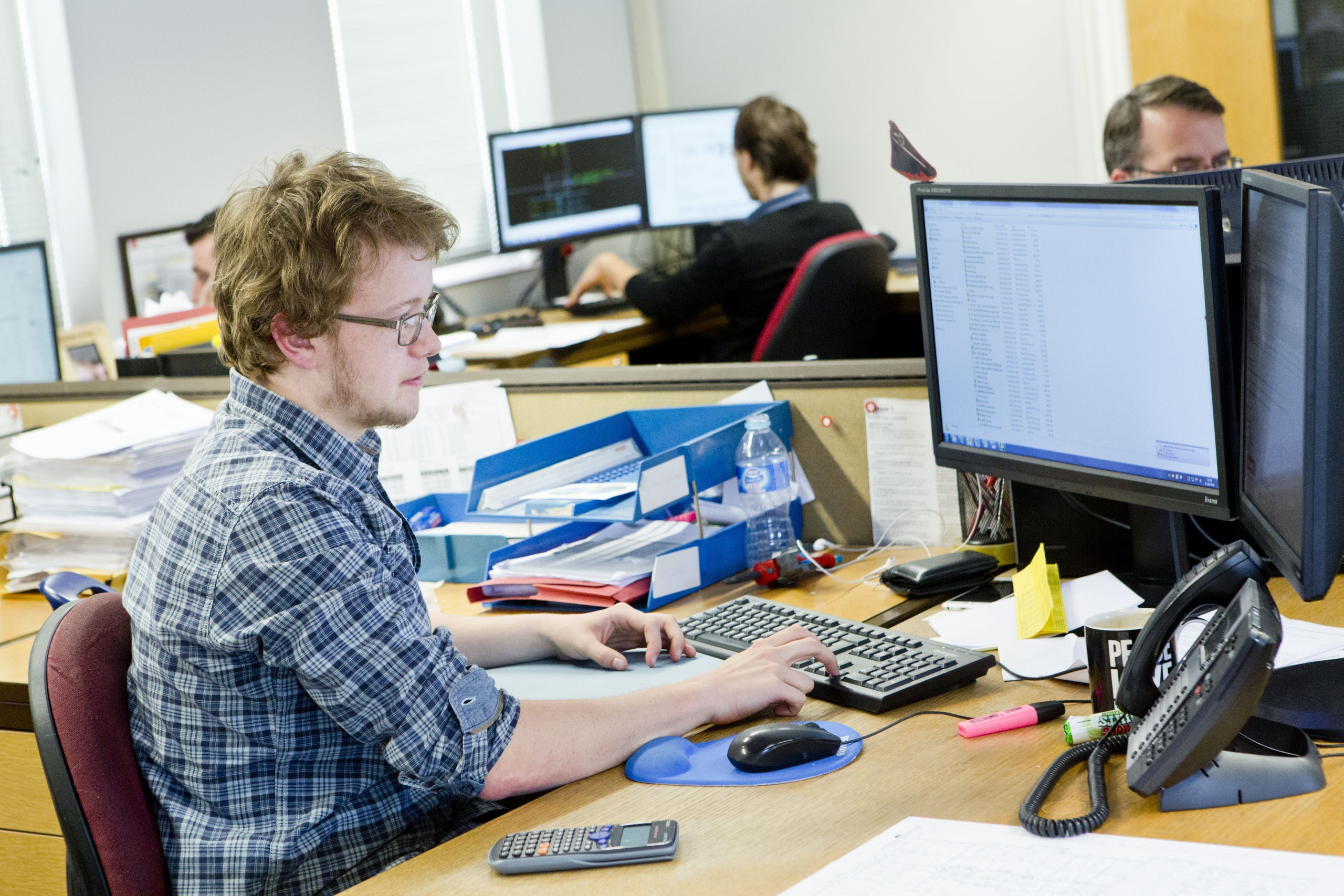 A designer sits at a computer reviewing files. Others are doing similar in the background.