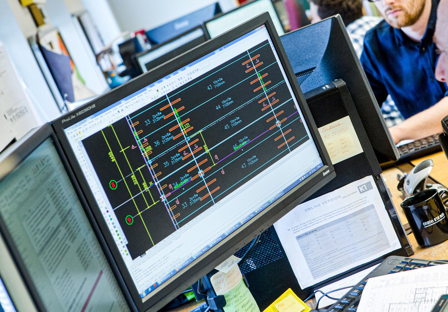 Workers in design office sit at computers with auto cad drawings open on screen