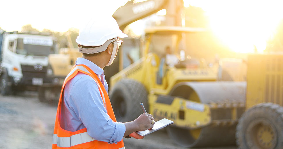 A construction worker writes on a clipboard whilst looking at machinery in the distance