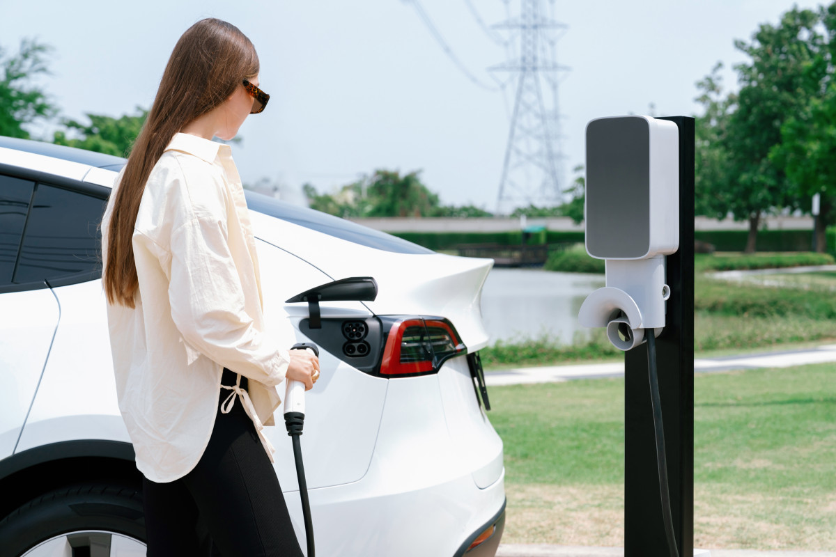A woman stands in front of a white car holding an EV charging cable